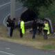 SUV flipped onto its side on the grassy roadside as responders and police stand nearby on a curve in the road.
