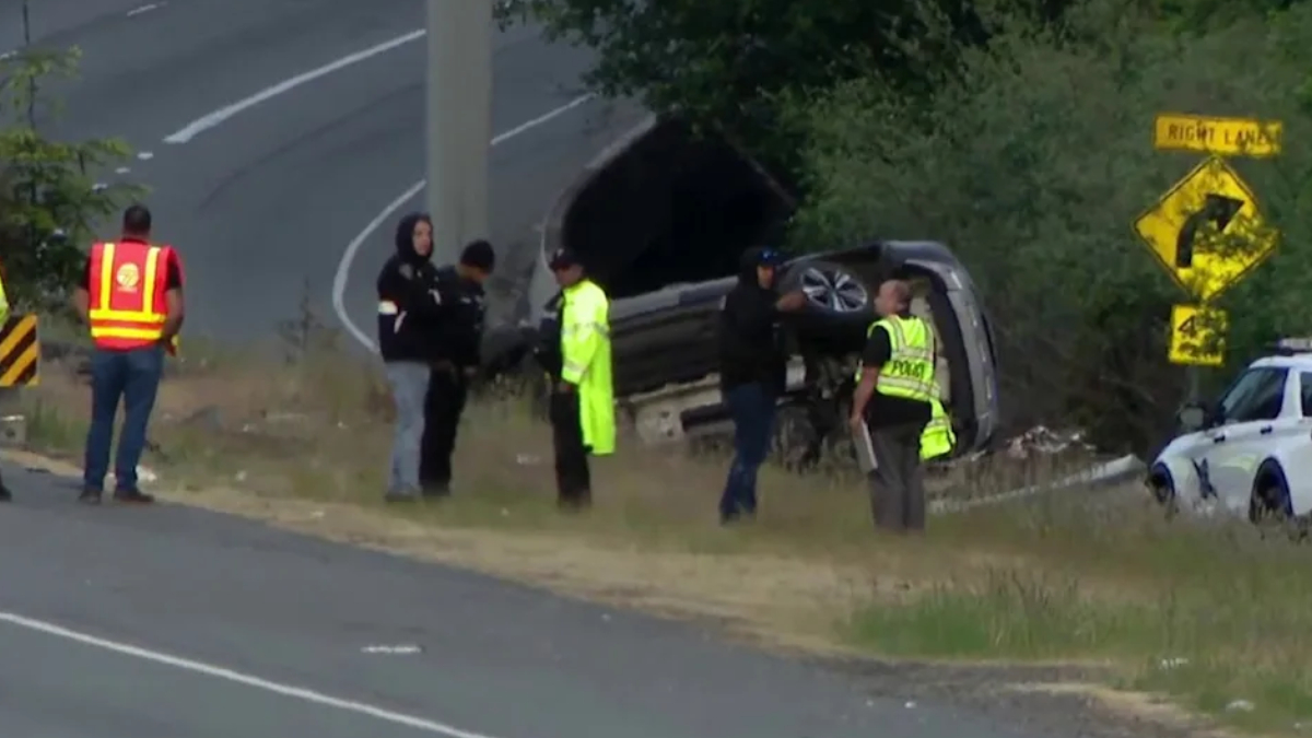 SUV flipped onto its side on the grassy roadside as responders and police stand nearby on a curve in the road.