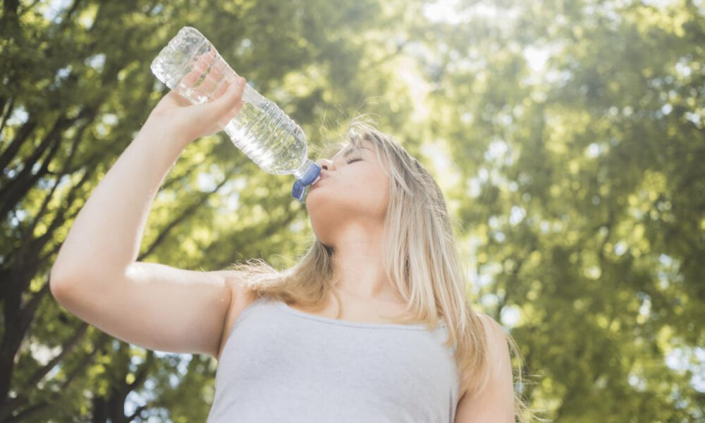 Young woman drinking water from a large plastic bottle outdoors in a sunny park with green trees behind her