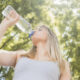 Young woman drinking water from a large plastic bottle outdoors in a sunny park with green trees behind her