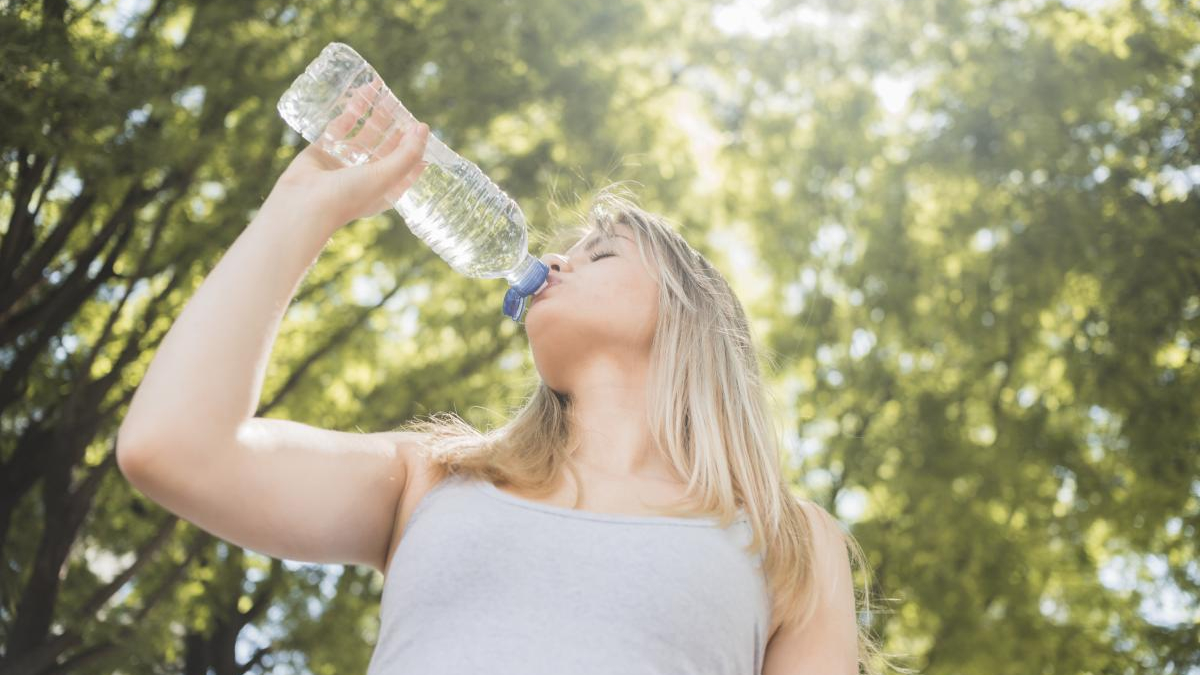 Young woman drinking water from a large plastic bottle outdoors in a sunny park with green trees behind her