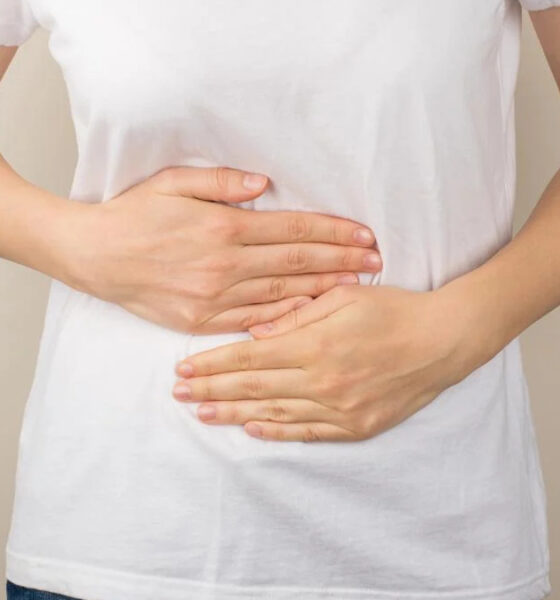 Person in a white shirt cups their abdomen with both hands, suggesting stomach pain or cramps.
