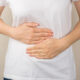 Person in a white shirt cups their abdomen with both hands, suggesting stomach pain or cramps.