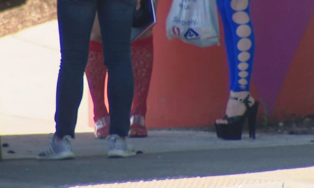Three pedestrians walk on a sidewalk beside a colorful mural; focus on legs and footwear: sneakers, red sandals, and black platform heels with blue patterned leggings (from behind).
