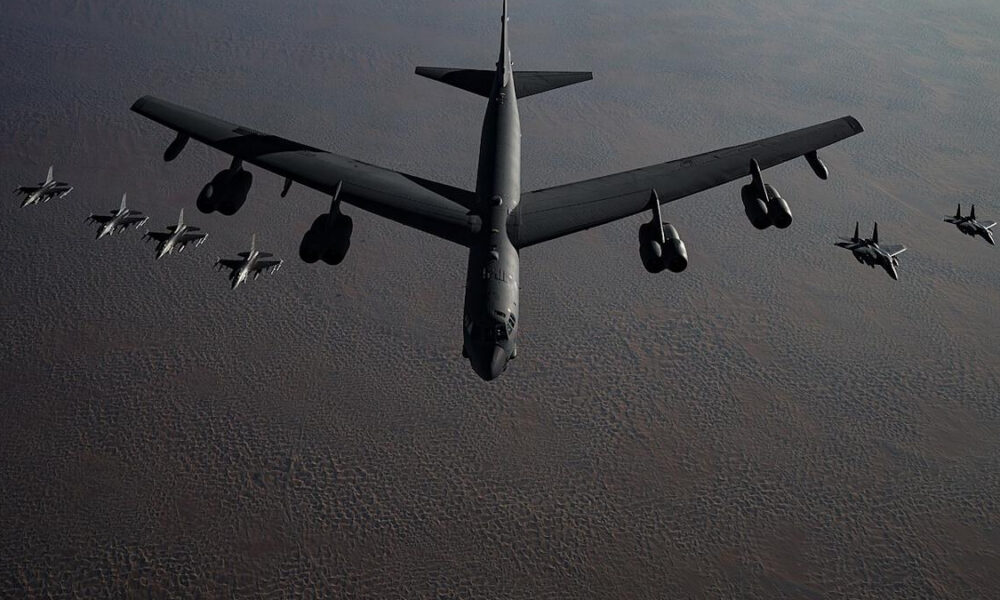 A large gray air tanker refueling aircraft with multiple fighter jets in formation above a desert-like sky and terrain on the horizon.
