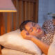 Person sleeping on a white pillow in bed with a warm bedside lamp nearby.