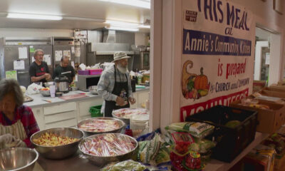Volunteers in a kitchen preparing meals, with large bowls of chopped ingredients on the counter and a banner reading 'His Meal at Annie's Community Kitchen' in the background.