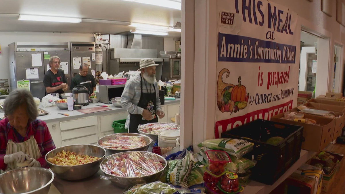 Volunteers in a kitchen preparing meals, with large bowls of chopped ingredients on the counter and a banner reading 'His Meal at Annie's Community Kitchen' in the background.