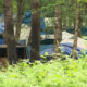 Cluster of blue and gray tents pitched among tall trees in a leafy forest campsite, with a small wooden shelter in the foreground.