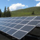 Rows of blue solar panels tilted on metal racks in a grassy field under a bright blue sky with trees nearby.
