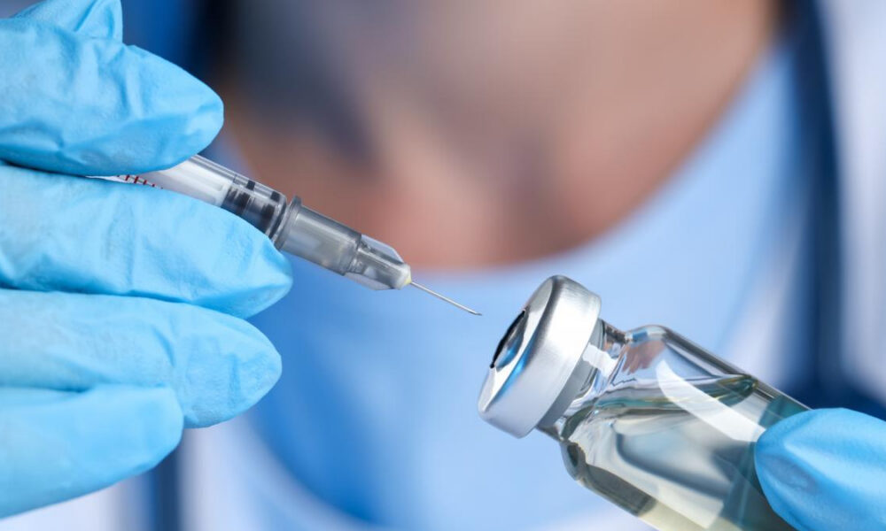 Close-up of a syringe being filled from a vial, hands wearing blue medical gloves visible in the foreground.