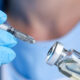 Close-up of a syringe being filled from a vial, hands wearing blue medical gloves visible in the foreground.