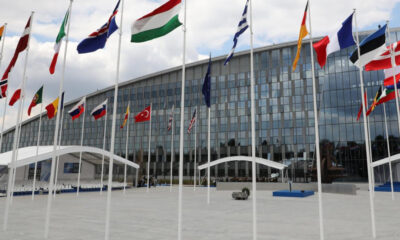 Modern glass-fronted building with many international flags on tall poles in a wide plaza.