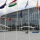 Modern glass-fronted building with many international flags on tall poles in a wide plaza.