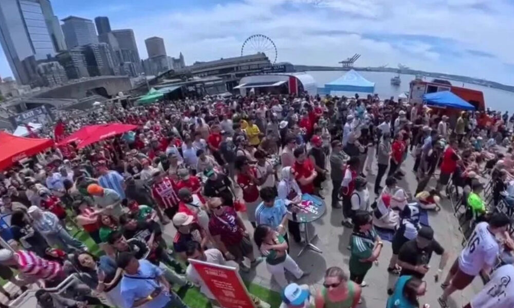Large crowd at an outdoor festival on a waterfront promenade with red and blue tents and a city skyline in the background.