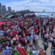 Large crowd at an outdoor festival on a waterfront promenade with red and blue tents and a city skyline in the background.