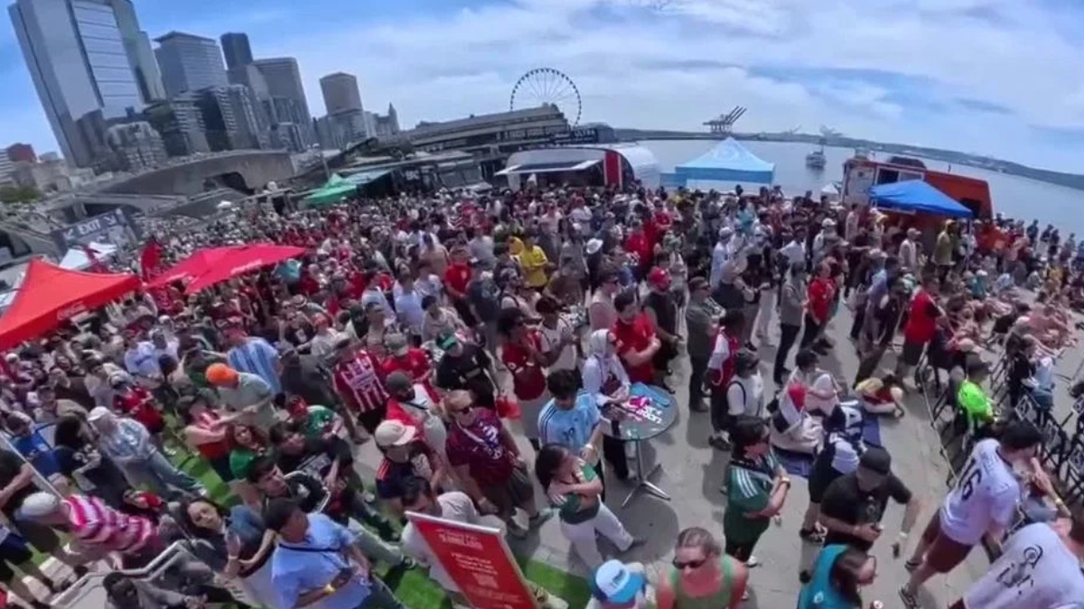 Large crowd at an outdoor festival on a waterfront promenade with red and blue tents and a city skyline in the background.
