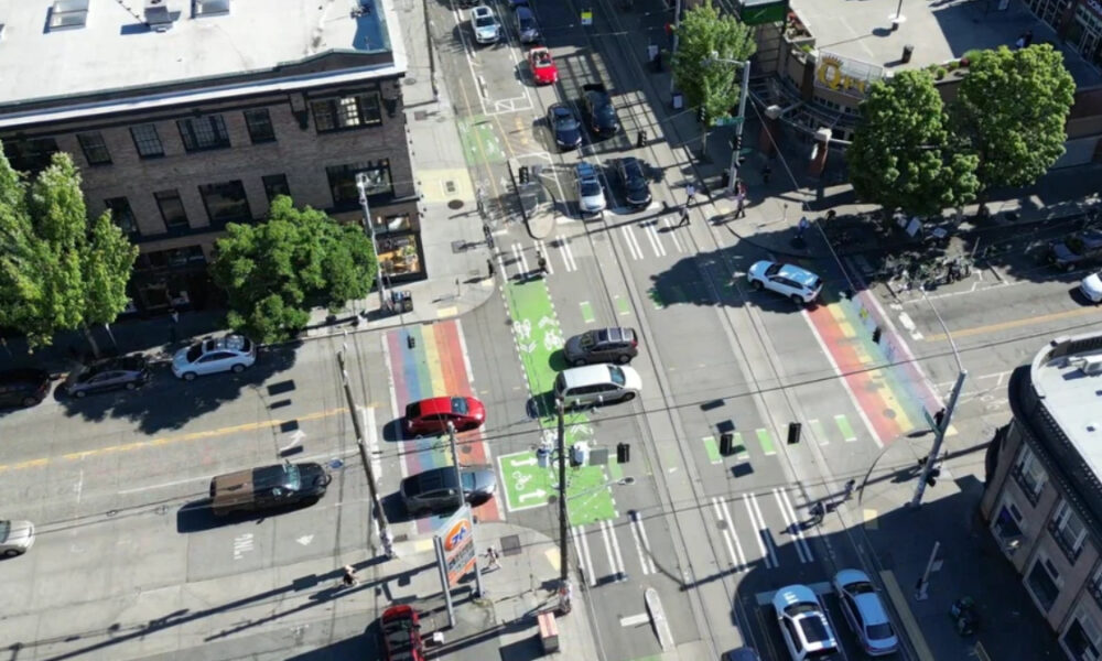 Aerial view of a city intersection with rainbow bike lanes and crosswalks, with cars and pedestrians nearby.