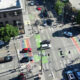 Aerial view of a city intersection with rainbow bike lanes and crosswalks, with cars and pedestrians nearby.