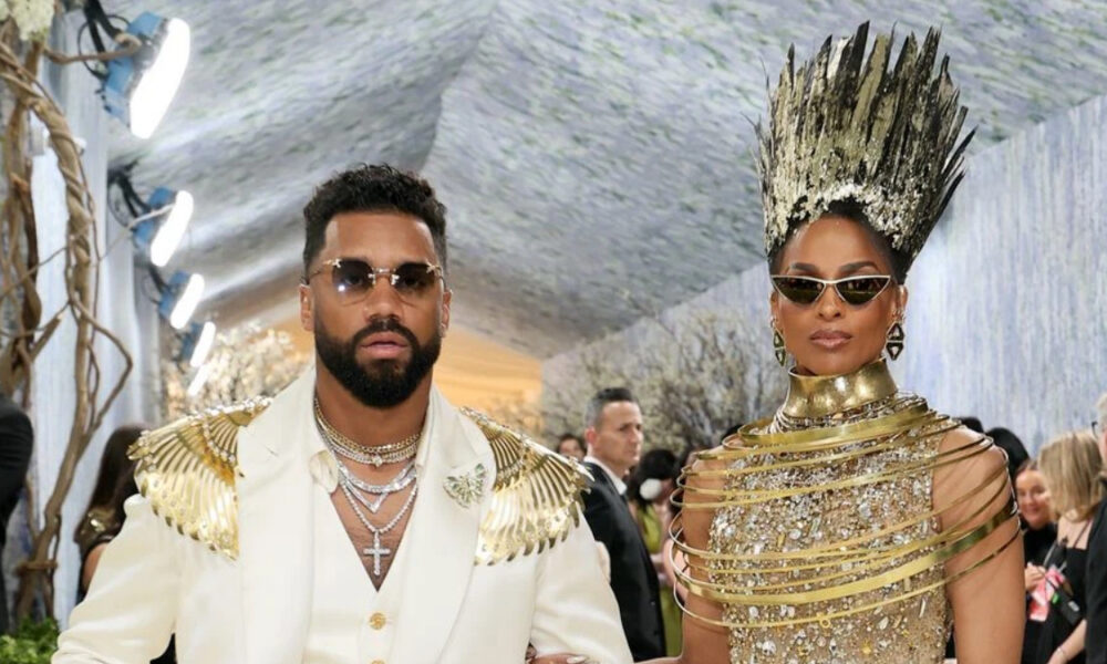 Man in a white suit with gold winged shoulder epaulets and layered chains, wearing sunglasses, stands beside a woman in a gold, cage-like gown and tall spiked crown at a gala.