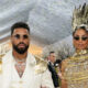 Man in a white suit with gold winged shoulder epaulets and layered chains, wearing sunglasses, stands beside a woman in a gold, cage-like gown and tall spiked crown at a gala.