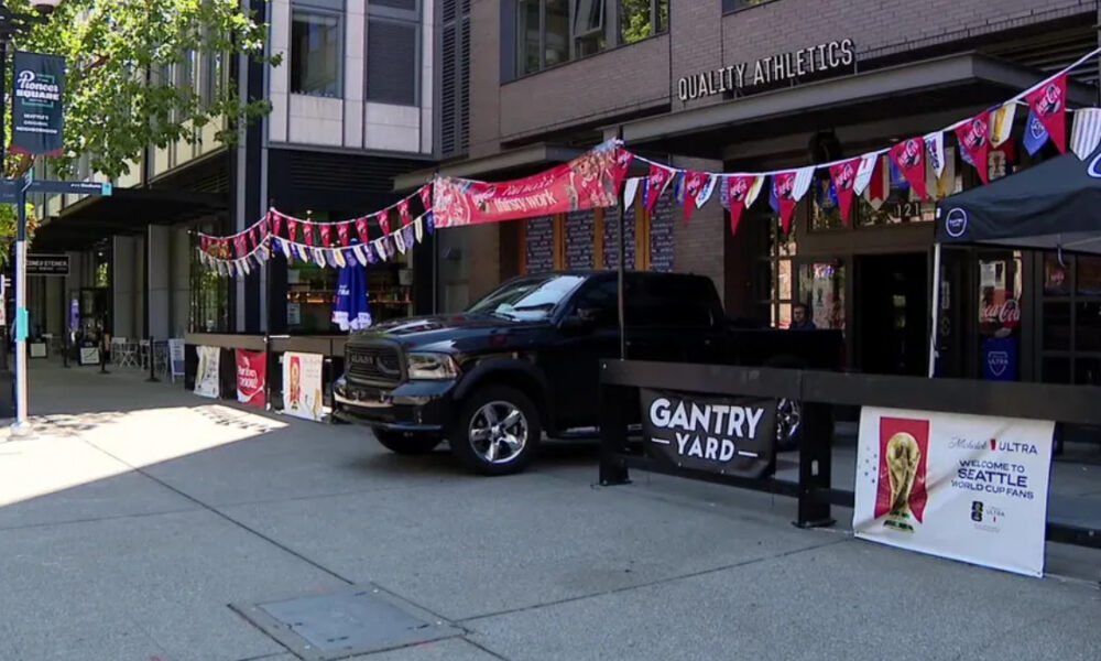 Black pickup truck parked on a city sidewalk next to a fenced event area called Gantry Yard, with colorful pennant banners overhead.