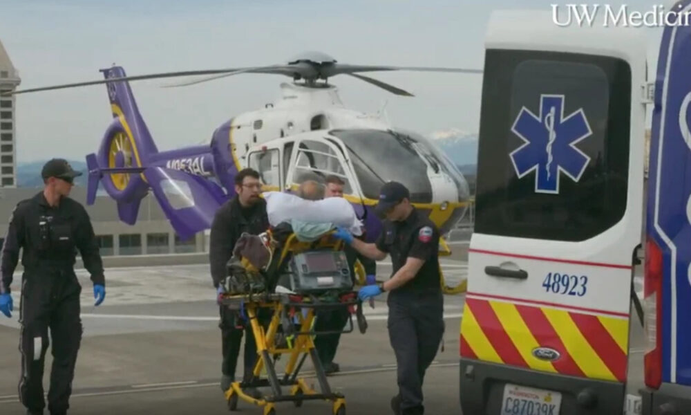 Emergency medical team transfers a patient on a stretcher toward an ambulance on a rooftop helipad beside a white-and-yellow helicopter (UW Medicine logo visible).