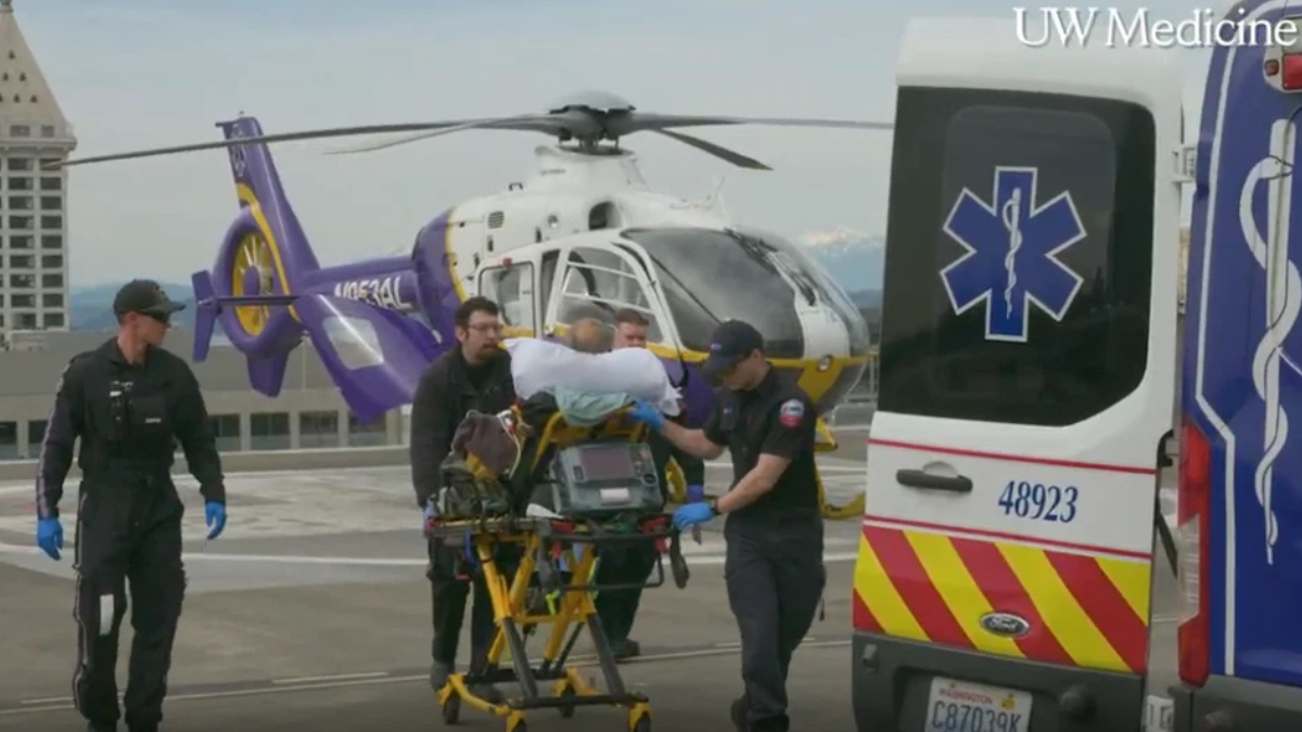 Emergency medical team transfers a patient on a stretcher toward an ambulance on a rooftop helipad beside a white-and-yellow helicopter (UW Medicine logo visible).