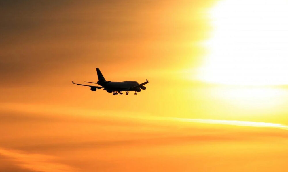 Silhouette of a commercial airliner with landing gear down, flying left to right against a bright orange sunset sky