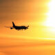 Silhouette of a commercial airliner with landing gear down, flying left to right against a bright orange sunset sky