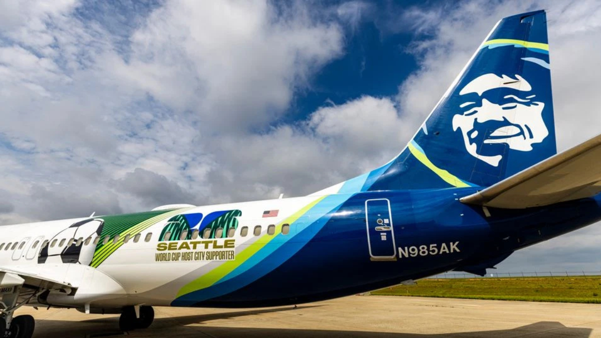 Commercial jet on a tarmac with a colorful Seattle World Cup host city supporter livery and a blue tail featuring a bold white graphic, registration N985AK.