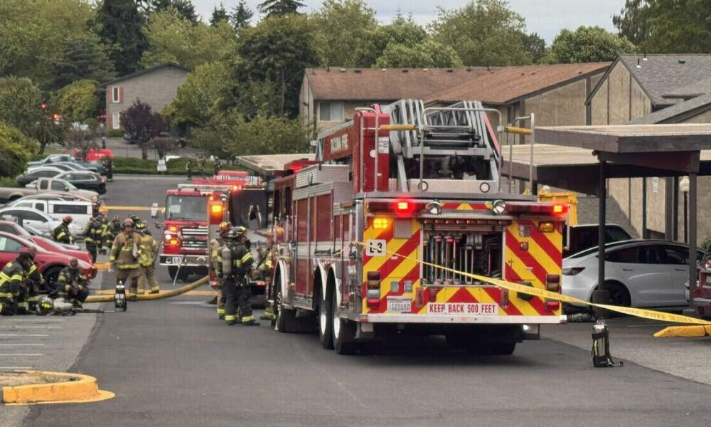 Emergency responders in full turnout gear pull hoses from a red fire truck at a suburban parking lot. 2) Fire department conducts incident with two trucks, hoses deployed, and caution tape across the area. 3) Firefighters kneel and prep equipment near a fire engine as others stand by. 4) Residential complex with beige buildings where emergency services are responding. 5) Back of fire truck showing chevrons and equipment, with 'KEEP BACK 500 FEET' sign visible.