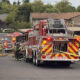 Emergency responders in full turnout gear pull hoses from a red fire truck at a suburban parking lot. 2) Fire department conducts incident with two trucks, hoses deployed, and caution tape across the area. 3) Firefighters kneel and prep equipment near a fire engine as others stand by. 4) Residential complex with beige buildings where emergency services are responding. 5) Back of fire truck showing chevrons and equipment, with 'KEEP BACK 500 FEET' sign visible.