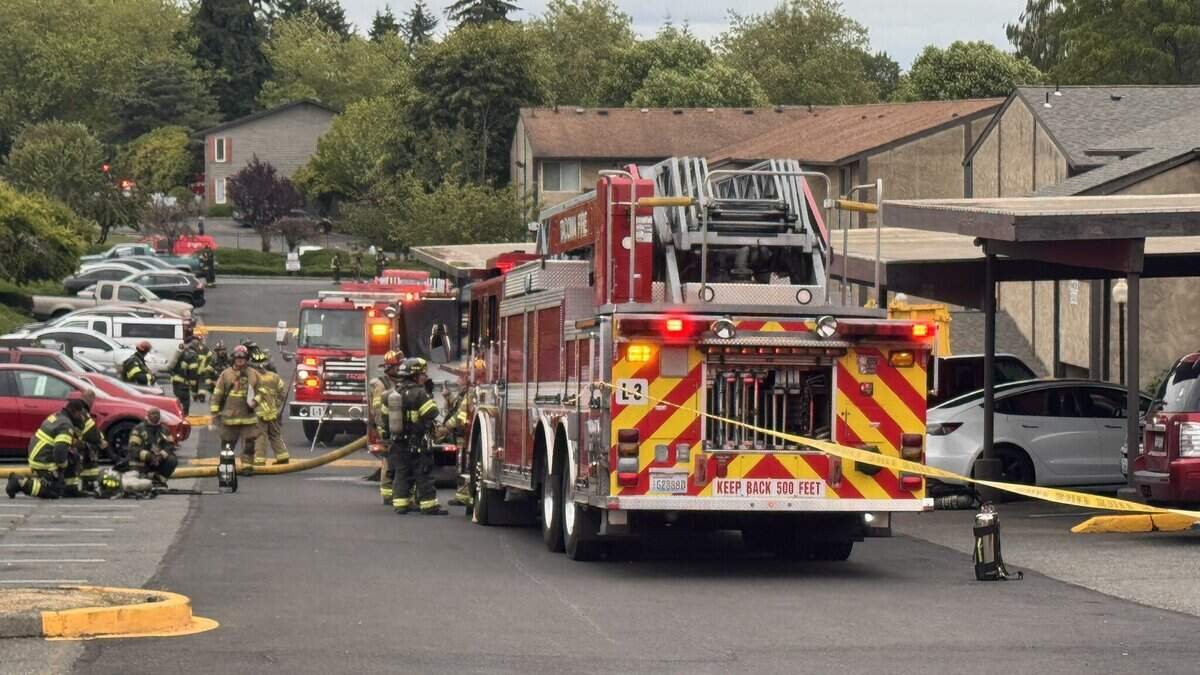 Emergency responders in full turnout gear pull hoses from a red fire truck at a suburban parking lot. 2) Fire department conducts incident with two trucks, hoses deployed, and caution tape across the area. 3) Firefighters kneel and prep equipment near a fire engine as others stand by. 4) Residential complex with beige buildings where emergency services are responding. 5) Back of fire truck showing chevrons and equipment, with 'KEEP BACK 500 FEET' sign visible.