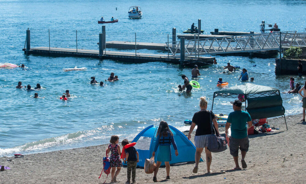 Family of five walks toward a blue pop-up tent on a sandy beach while swimmers enjoy the calm water nearby and a pier extends into the sea in the background.