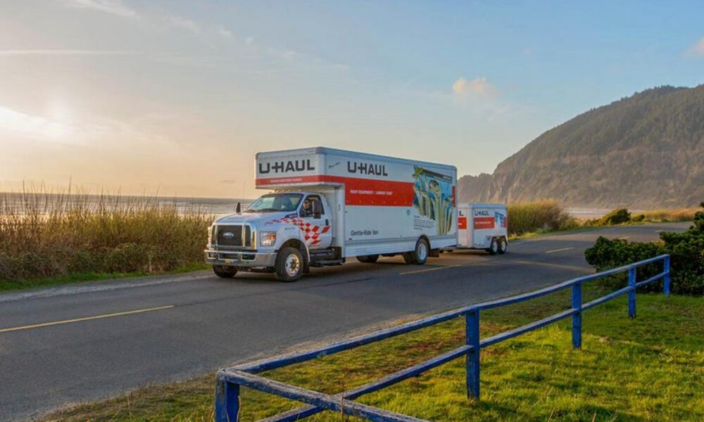 White U-Haul moving truck with red stripe and trailer on a coastal road at sunset.