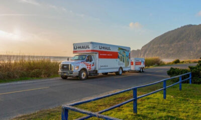White U-Haul moving truck with red stripe and trailer on a coastal road at sunset.