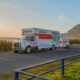 White U-Haul moving truck with red stripe and trailer on a coastal road at sunset.