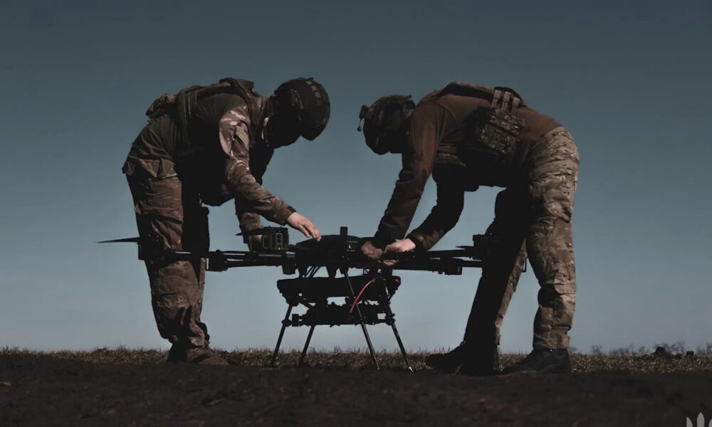 Two soldiers in camouflage gear assemble a large quadcopter drone on a dirt field under a clear sky.