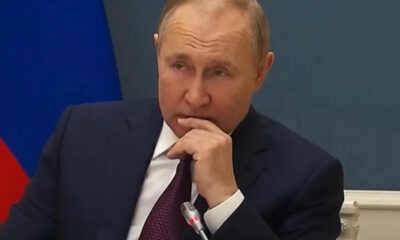 Man in a dark suit and maroon tie sits at a table, resting his chin on his hand while speaking into a microphone, with a blue wall and flag in the background.
