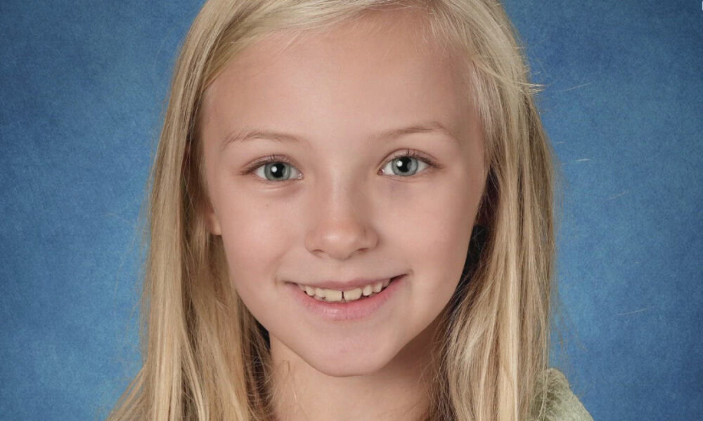 Smiling young girl with long blonde hair in front of a blue textured backdrop, facing the camera.