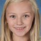 Smiling young girl with long blonde hair in front of a blue textured backdrop, facing the camera.