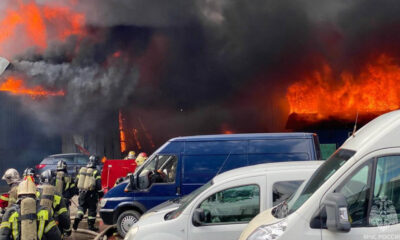 Firefighters in full gear battle a large warehouse blaze as thick black smoke and bright flames billow from the roof, with parked vans in the foreground.