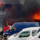 Firefighters in full gear battle a large warehouse blaze as thick black smoke and bright flames billow from the roof, with parked vans in the foreground.