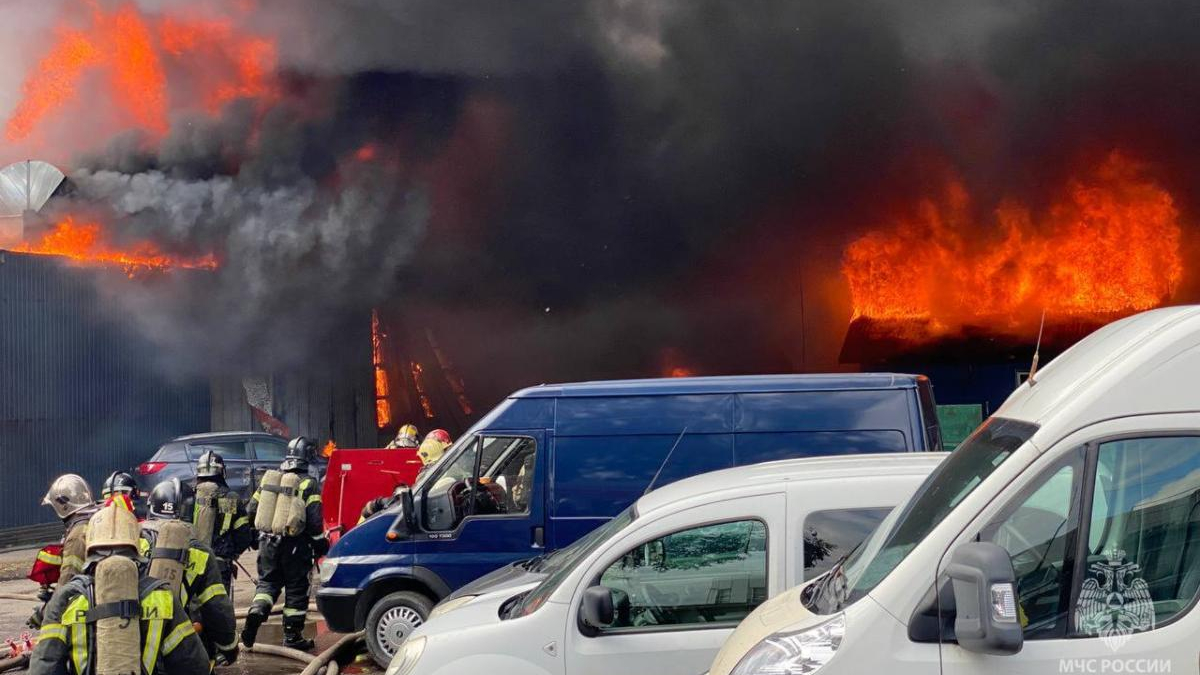 Firefighters in full gear battle a large warehouse blaze as thick black smoke and bright flames billow from the roof, with parked vans in the foreground.