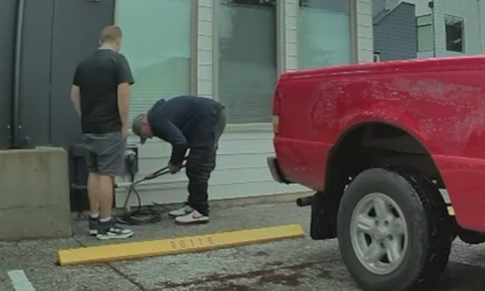 Two men work near a gray building with equipment on the ground beside a red pickup truck in a parking lot, possibly setting up or inspecting gear.