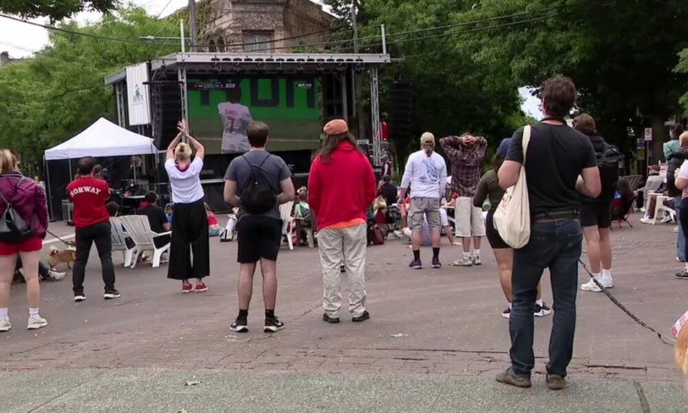 Outdoor community event with a stage and crowd facing it; people stand and sit on white chairs in a park-like plaza.
