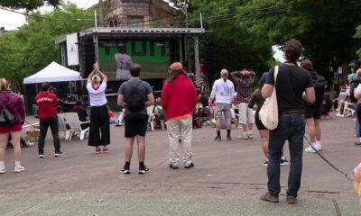 Outdoor community event with a stage and crowd facing it; people stand and sit on white chairs in a park-like plaza.