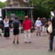 Outdoor community event with a stage and crowd facing it; people stand and sit on white chairs in a park-like plaza.