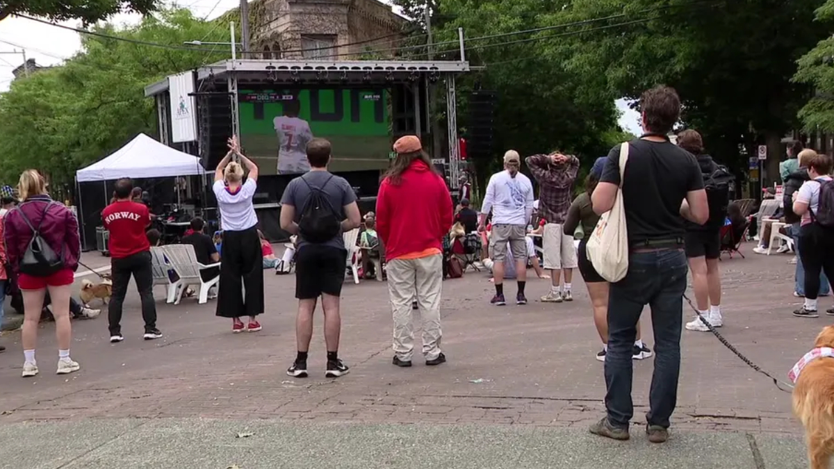 Outdoor community event with a stage and crowd facing it; people stand and sit on white chairs in a park-like plaza.